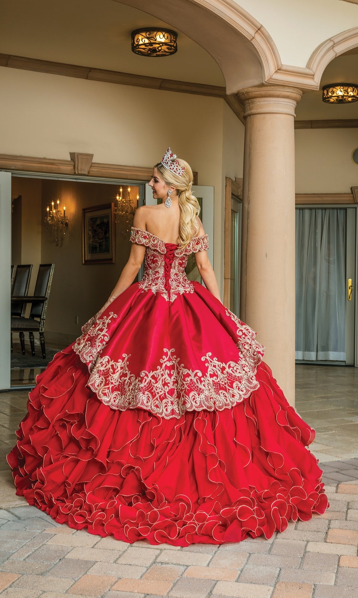 A woman in the Quinceanera Dress 1599 by Dancing Queen, a ruffle-layered off-the-shoulder ball gown with gold embroidery, stands outside facing away, her blonde curls flowing beneath a tiara.